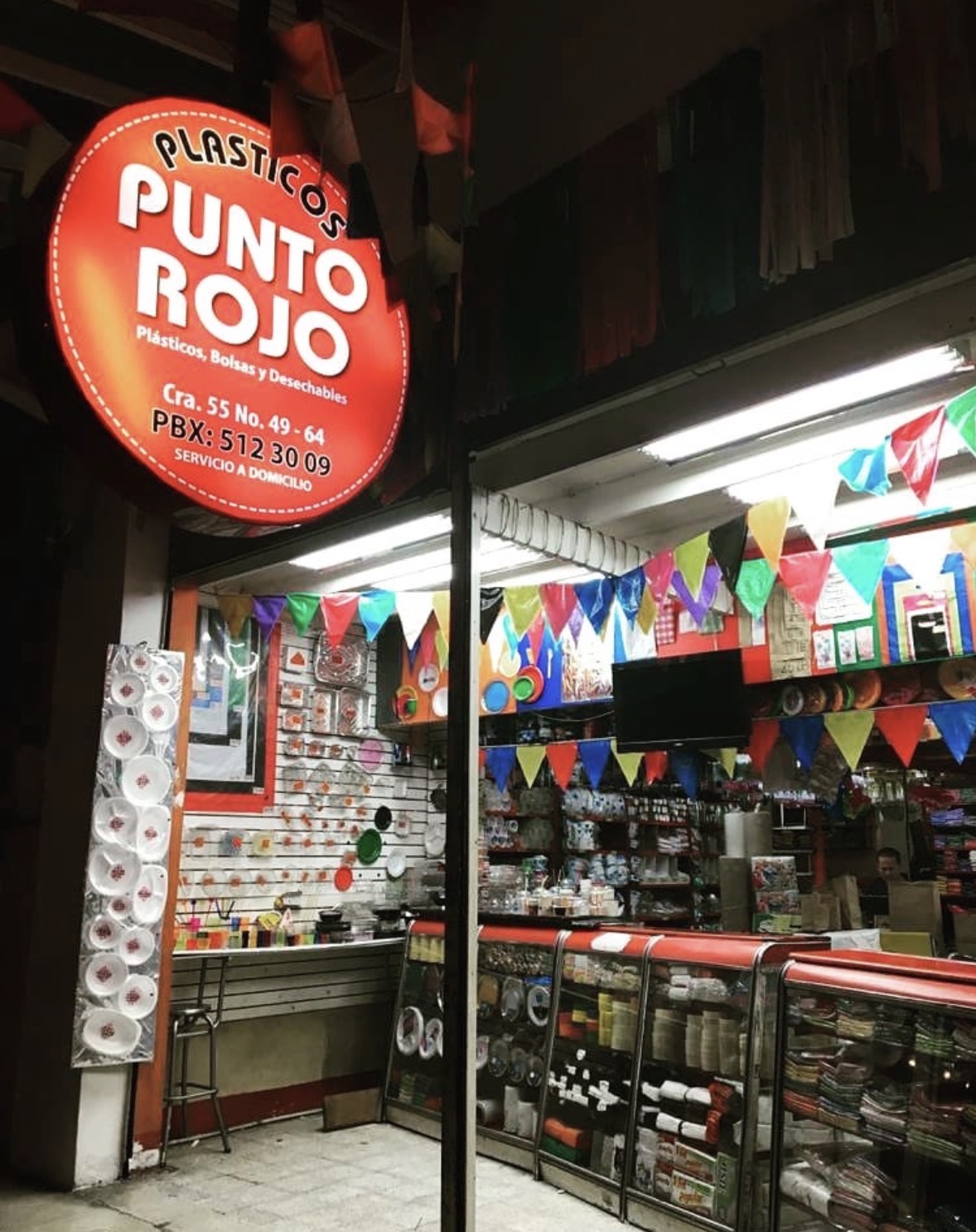 La fachada de Plásticos Punto Rojo en la Carrera 55: el aviso rojo con el nombre y la dirección, y adentro los estantes llenos de vasos, platos y desechables.